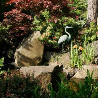 A serene garden scene featuring a heron sculpture surrounded by lush foliage and large rocks.