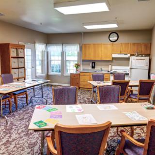 A well-lit office space with tables and chairs, featuring a kitchenette along one wall.