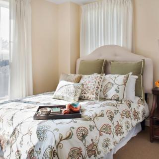 A cozy bedroom with a neatly made bed featuring floral-patterned bedding,
a headboard, and decorative pillows, situated near a window with sheer
curtains. A tray with items is placed on the bed.