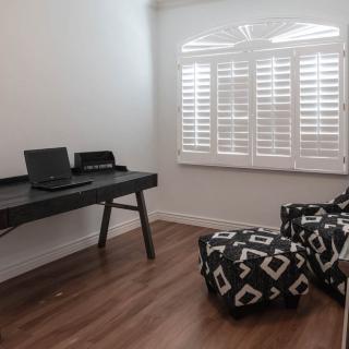 A minimalist home office with a laptop on a desk, patterned ottomans, and a window with shutters.