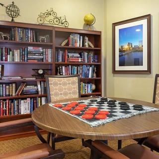 A cozy game room with a checkers board set up on the table, surrounded by bookshelves and artwork on the wall.