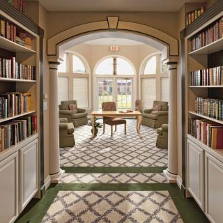 A cozy reading area with bookshelves, comfortable chairs, and a table, illuminated by natural light through large windows.