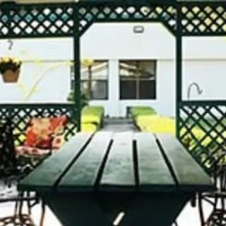 An outdoor patio area featuring a long wooden table, surrounded by chairs and lattice fencing with hanging plants.