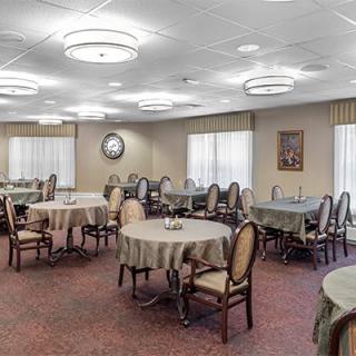 A well-lit dining area with multiple round tables and upholstered chairs arranged on a carpeted floor.