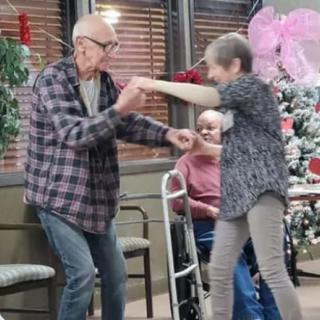 An elderly couple joyfully dances together indoors, surrounded by festive decorations.