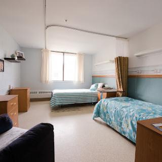 A well-lit dormitory room featuring two neatly made beds, a desk, and ample storage space.