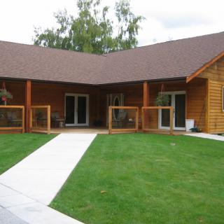 A charming wooden house with a lush green lawn and a concrete pathway leading to the entrance.