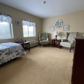 A cozy bedroom with floral decor, featuring two armchairs, a bed, and windows allowing natural light.