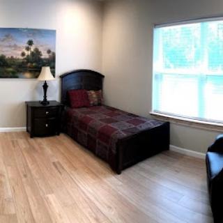 A serene and minimalist bedroom featuring a single bed, a nightstand with a lamp, and a large window that lets in natural light.