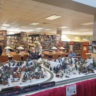 A festive miniature train and village display set up in a library, surrounded by bookshelves.