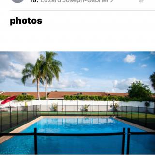 A backyard pool surrounded by a safety fence with palm trees and a clear blue sky in the background.