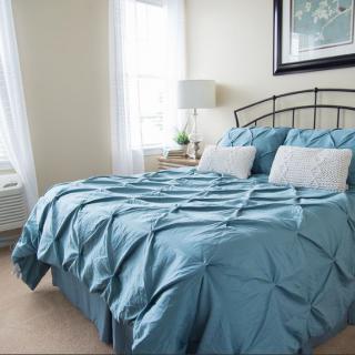A cozy bedroom with a bed covered in blue bedding and pillows, featuring a lamp and natural light from the windows.