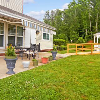 A backyard patio area with outdoor furniture, surrounded by a lawn and garden, featuring potted plants and a gazebo in the background.