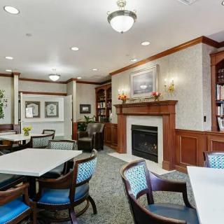 A cozy library interior featuring a fireplace, tables, chairs, and bookshelves.