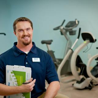 A fitness trainer posing in a gym with exercise equipment in the background.