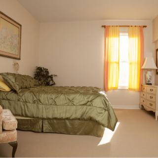 A cozy bedroom with warm lighting, featuring a bed with green bedding, a cushioned chair, and a dresser beside a window with orange curtains.