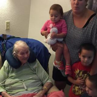 A family gathering with an elderly woman seated and surrounded by children and a caregiver.