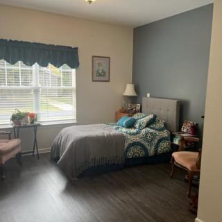 A cozy bedroom with a single bed, featuring a window with blue curtains, a lampshade on the bedside table, and chairs next to the bed.