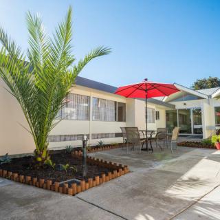 A sunny patio area featuring a red umbrella, outdoor furniture, and a palm tree beside a house.