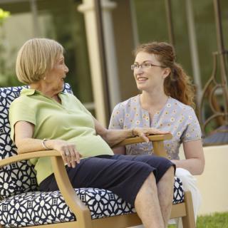An elderly woman sits in a chair while having a conversation with a young woman outside.