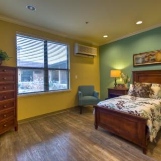 A cozy bedroom with a bed, dresser, and armchair, featuring warm lighting and a large window.