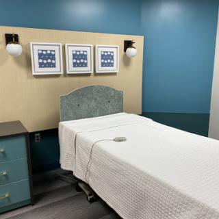A modern hospital patient room featuring a neatly made bed, wall art, and soothing blue tones.