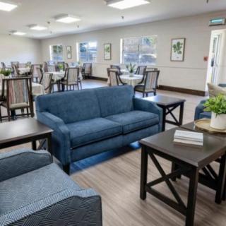 A spacious and well-lit living room with blue sofas and a dining area in the background.