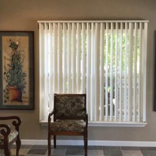 A room interior featuring a window with vertical blinds, flanked by framed artwork and a wooden chair.
