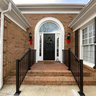 A welcoming brick house entrance with a black front door adorned with a wreath, flanked by decorative lights and handrails.