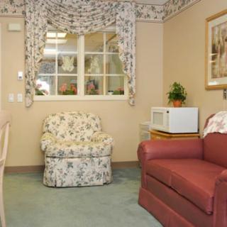 A cozy living room featuring a floral armchair and red sofa, with a microwave on a side table and decorative curtains framing a window.
