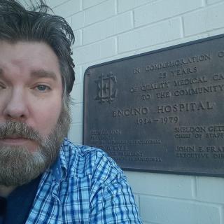 A man with a beard takes a selfie in front of a plaque on a brick wall.