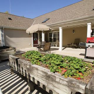 A sunny backyard patio featuring a raised garden bed, outdoor furniture, an umbrella, and a barbecue grill.