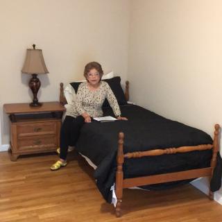 A woman sitting on a bed in a neatly arranged bedroom with wooden flooring.