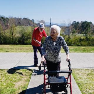 An elderly woman using a walker, accompanied by a man, enjoying a sunny day in the park.