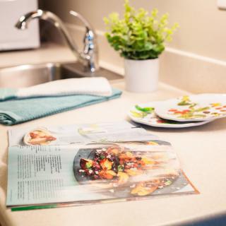 A kitchen countertop with an open recipe book, a small plant, and clean dishes near the sink.