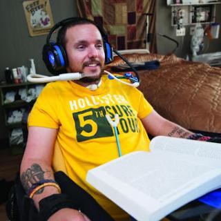 A man in a yellow shirt enjoys gaming using assistive technology while seated in a wheelchair.