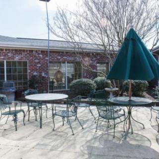 A sunny outdoor patio area with tables, chairs, and a large umbrella beside a brick building.