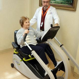 An elderly woman uses an exercise machine with the assistance of a healthcare professional during a rehabilitation session.
