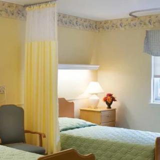 A hospital room with two beds separated by a curtain, featuring a table lamp and a window.