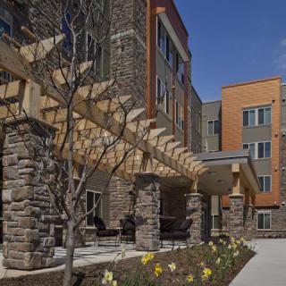 A modern residential building featuring a stone facade and a wooden pergola, surrounded by landscaped greenery.
