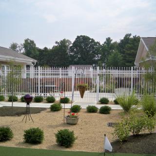A landscaped garden area with a white fence, various bushes, and plants set against a residential backdrop.