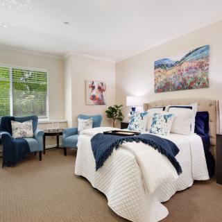 A cozy, modern bedroom featuring a neatly made bed with decorative pillows and throws, two plush armchairs by the window, and vibrant artwork adorning the walls.