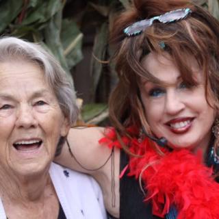 Two women smiling together outdoors, one wearing a red feather boa.