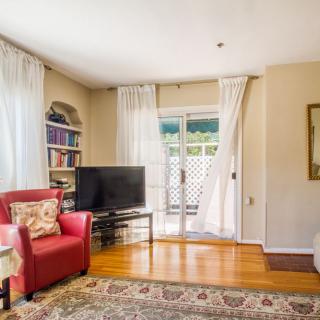 A cozy living room with a red armchair, bookshelves, and a TV sits atop a hardwood floor with a patterned rug, leading to a sliding door covered by sheer curtains.