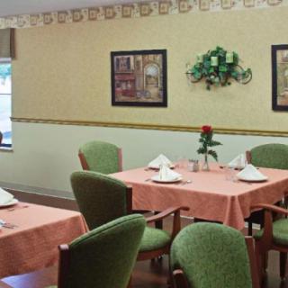 A cozy dining room with neatly set tables, featuring green chairs and floral centerpieces.