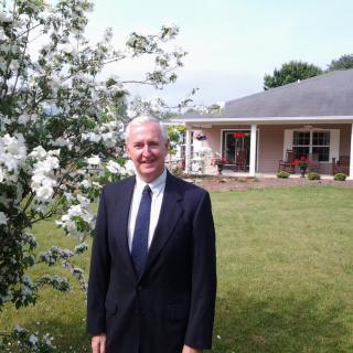 A man in a suit standing outside near flowering plants with a house in the background.