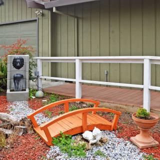 A small decorative garden featuring a mini wooden bridge, gravel pathway, planter, and water fountain next to a wooden deck with railing.