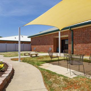 A sunny courtyard featuring a shade sail, patio furniture, and landscaped surroundings.