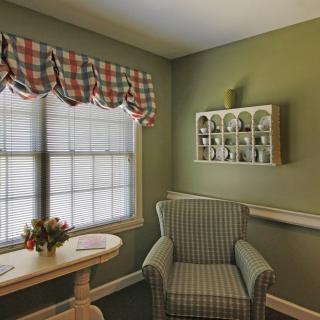 A cozy corner of a living room featuring a plaid-patterned armchair, small round table with a vase of flowers, and decorative shelves on the wall. A window with checkered curtains adds charm to the space.