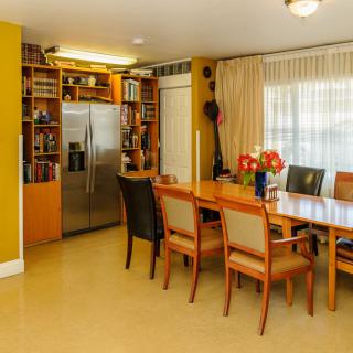 A well-lit dining area with a large table and chairs, adjacent to a kitchen space featuring a stainless steel refrigerator.